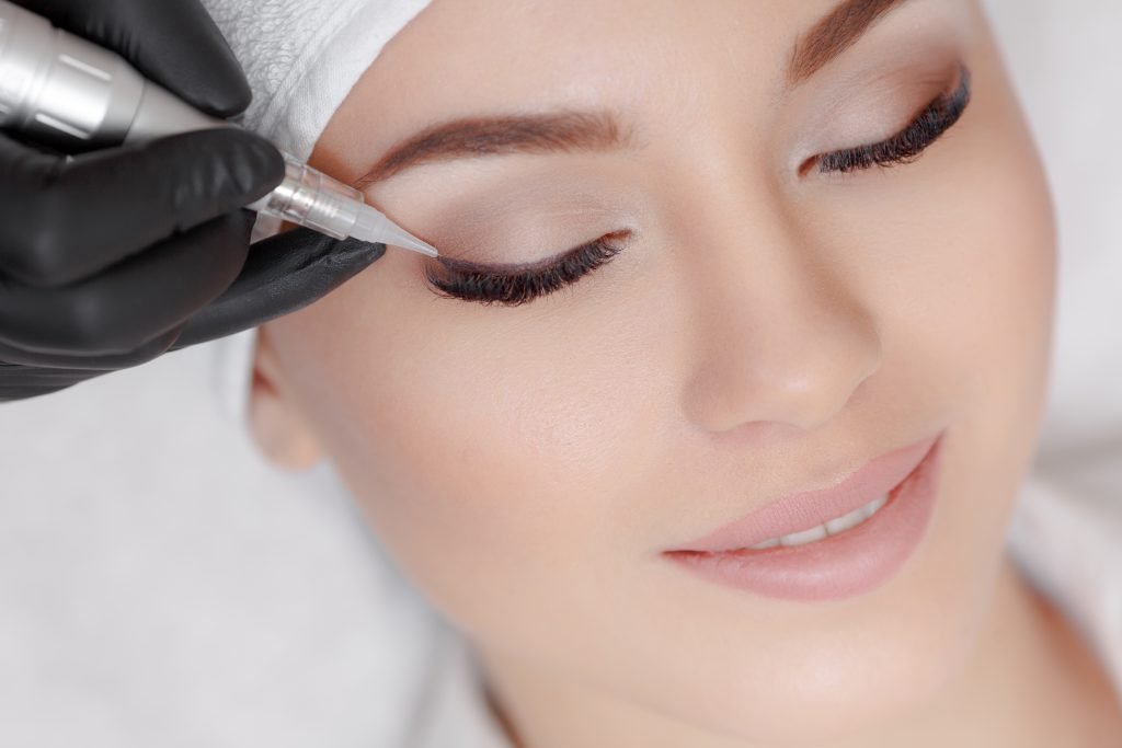 Close up of woman lying on couch at beauty salon with closed eyes and enjoying when cosmetologist making eyeliner permanent make up. Professional wearing black gloves and holding speciality tool.